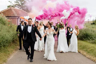Bride holding bouquet of flowers with wedding party holding pink, white and orange smoke flares. 