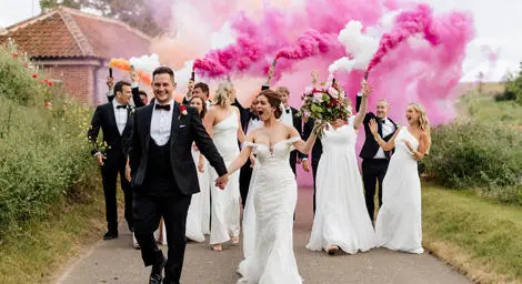 Bride holding bouquet of flowers with wedding party holding pink, white and orange smoke flares. 
