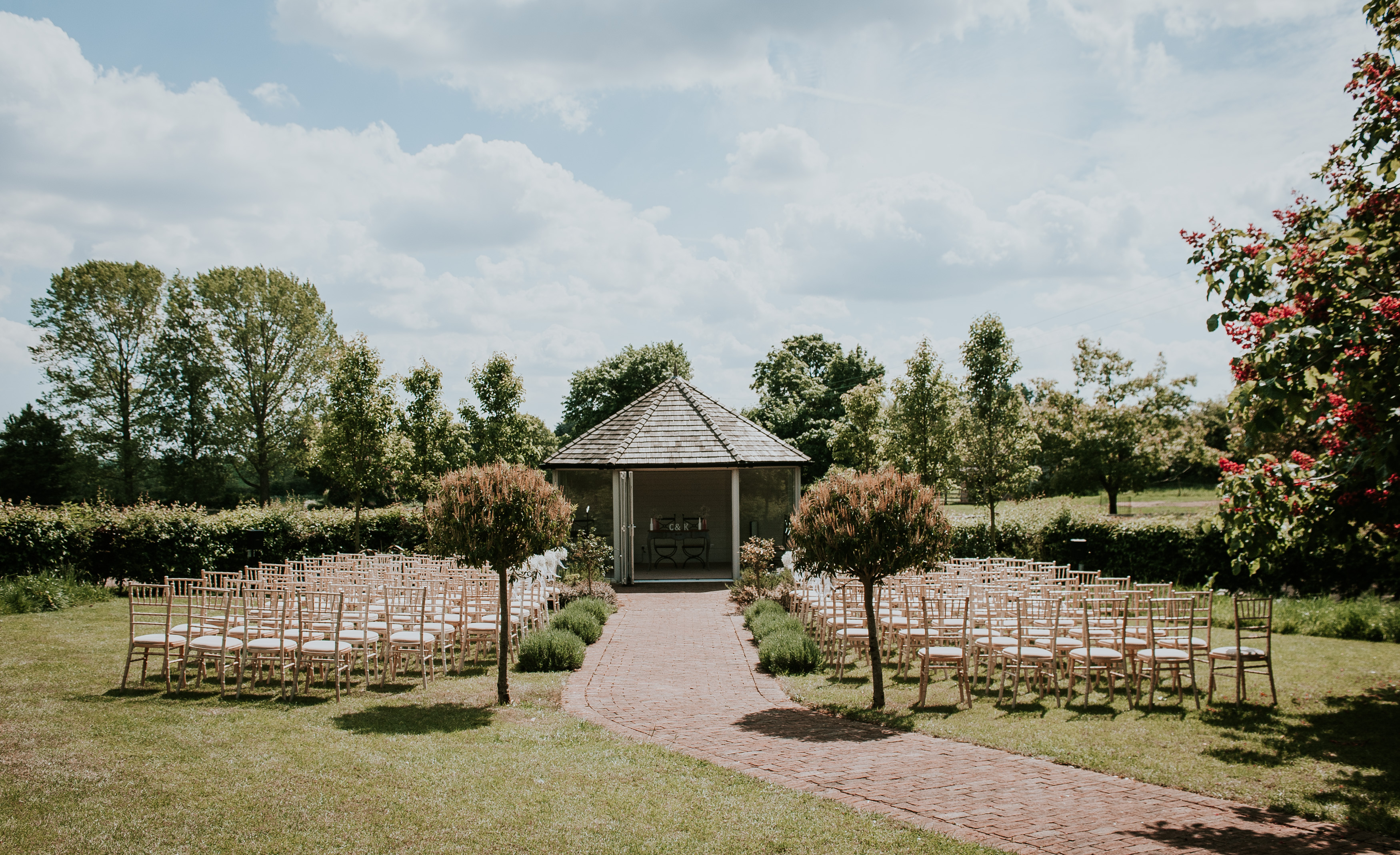 Paved path leading to bandstand with chairs either side of it. Trees and fields are in the background.