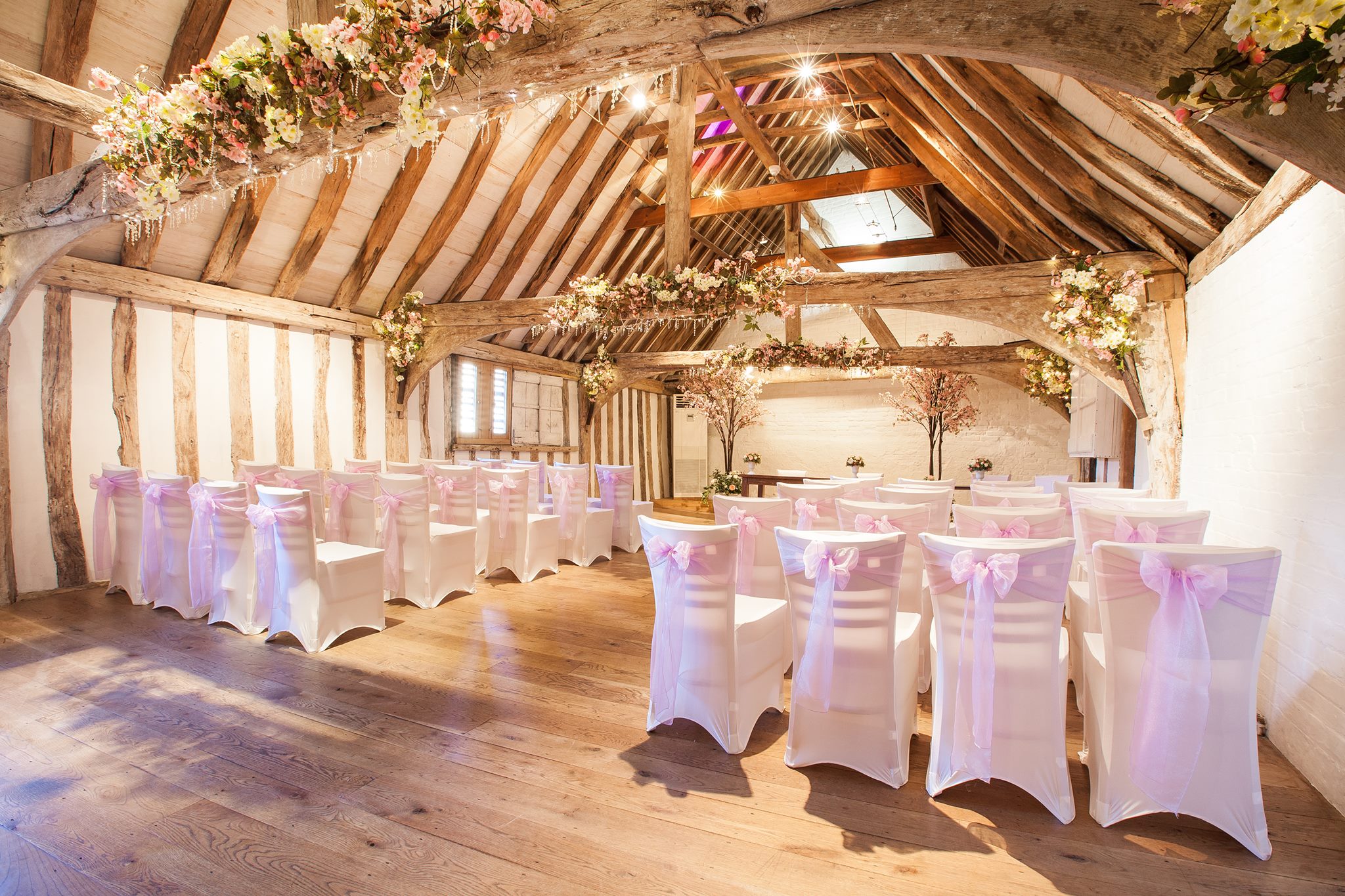 Pink ribbon tied into bows round white chairs in a room with wooden beams and flowers decorating it.