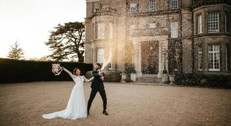 Bride and groom in front of grand mansion laughing and spraying champagne