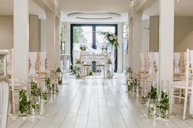 Chairs facing towards a flower arch. Flowers in glass vases and petals on the edge of the aisle.
