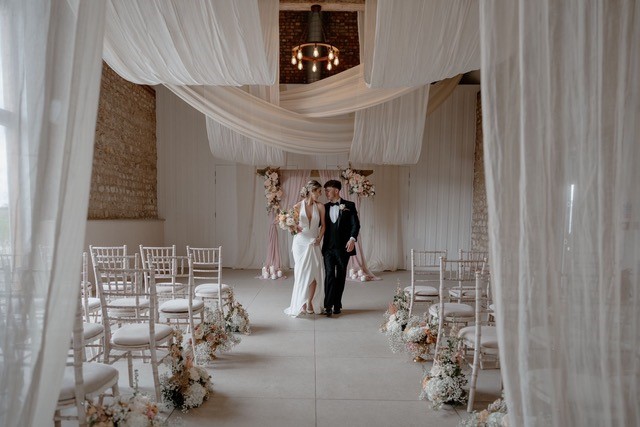 Bride and groom in beautifully decorated ceremony room with white chairs, white flowers and white drapery