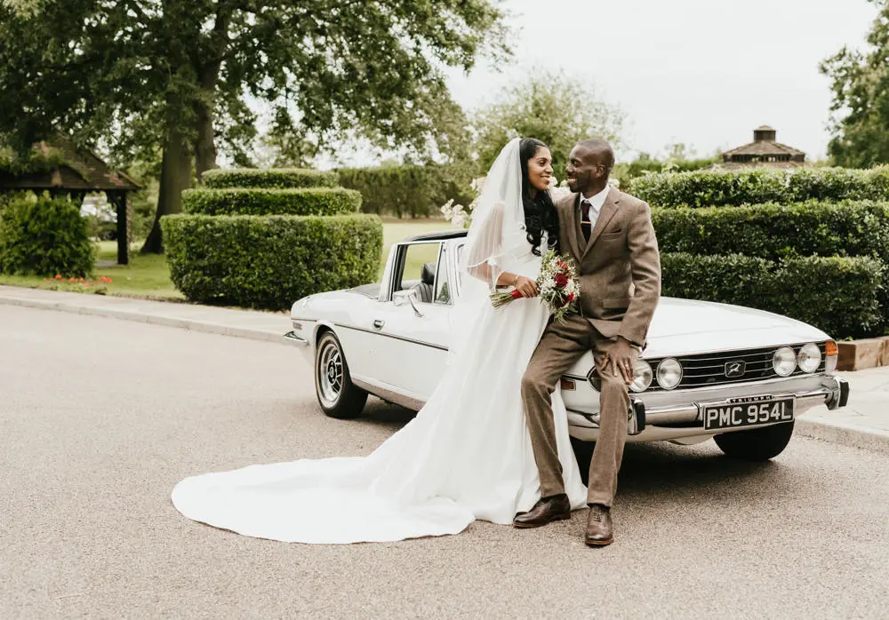 Bride and groom in parkland setting leaning against classic car