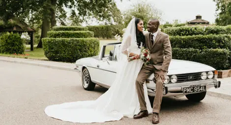 Bride and groom in parkland setting leaning against classic car