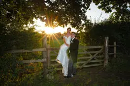 Bride sitting on countryside gate with groom in a morning suit next to her. 
