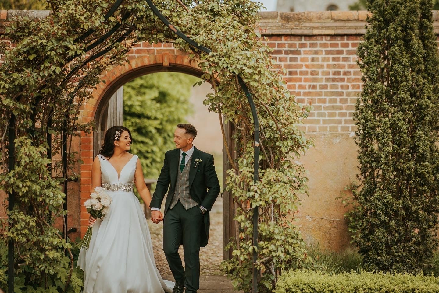 White bride and groom holding hands smiling at each other walking under a garden arch.