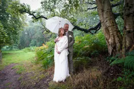 Groom holding white umbrella stood behind bride in the forest. Bride smiling at groom