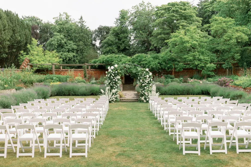 White chairs outside facing a flower arch, trees and a courtyard.