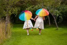 Two brides holding rainbow umbrellas and lifting feet to see rainbow sole on trainers. 