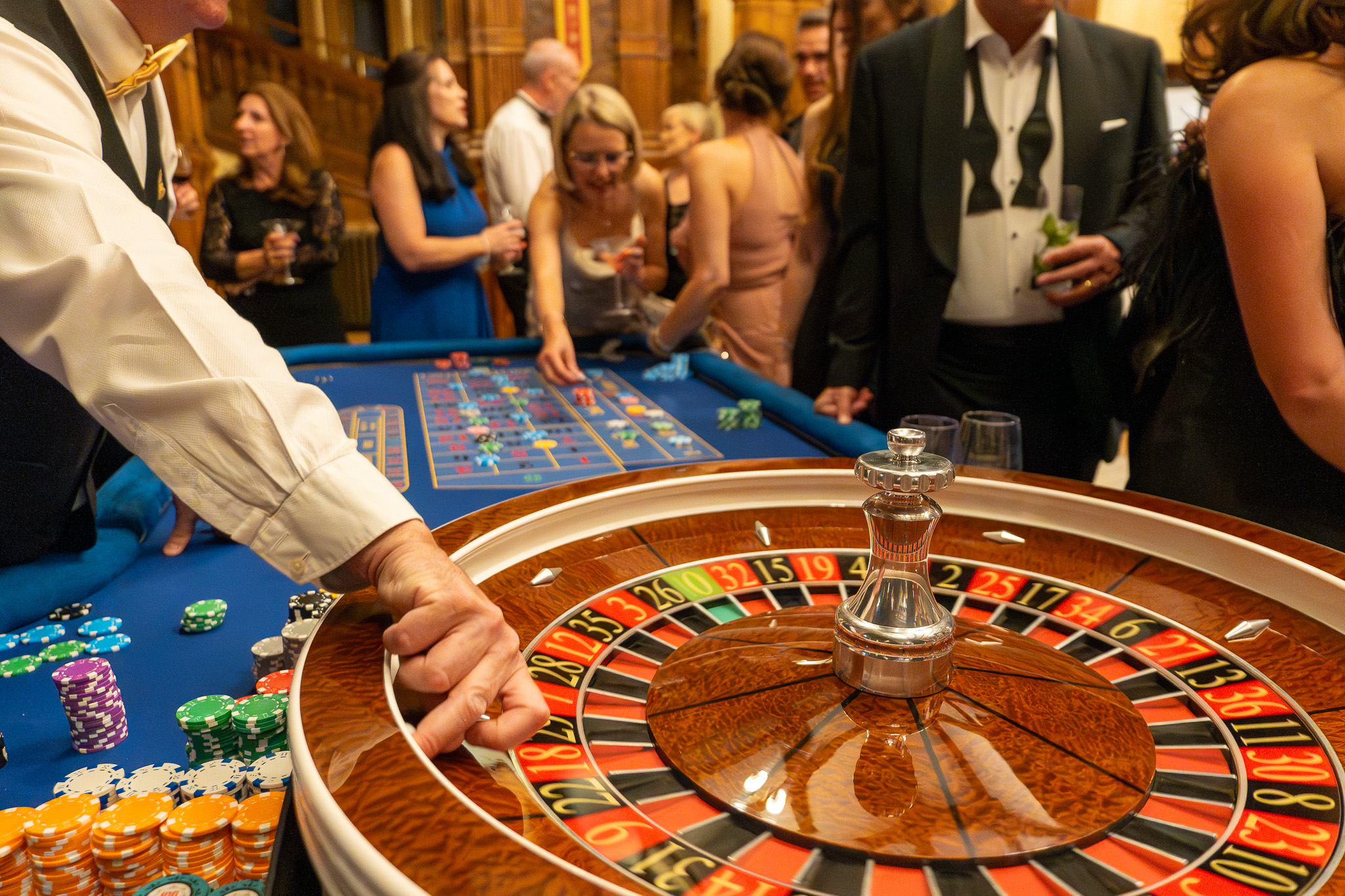 Roulette wheel with black tie guests in the background and showing part of croupiers arm releasing the ball
