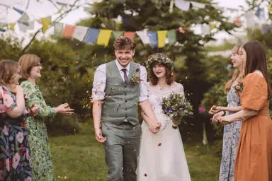 Bride wearing flower crown holding hands with groom with purple and yellow confetti thrown at them