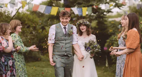 Bride wearing flower crown holding hands with groom with purple and yellow confetti thrown at them