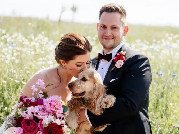 Bride kissing dog with groom in a meadow. 