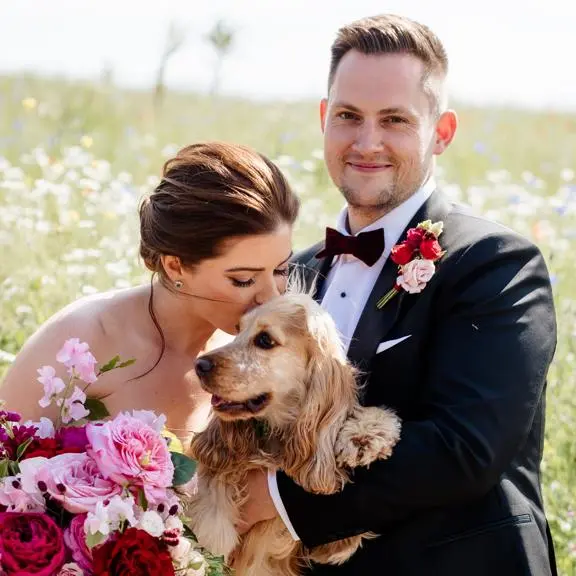 Bride kissing dog with groom in a meadow.