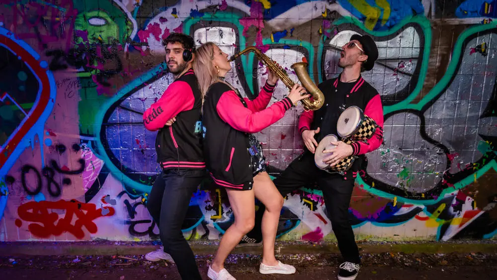 Three musicians playing saxophone and drums in front of graffiti wall