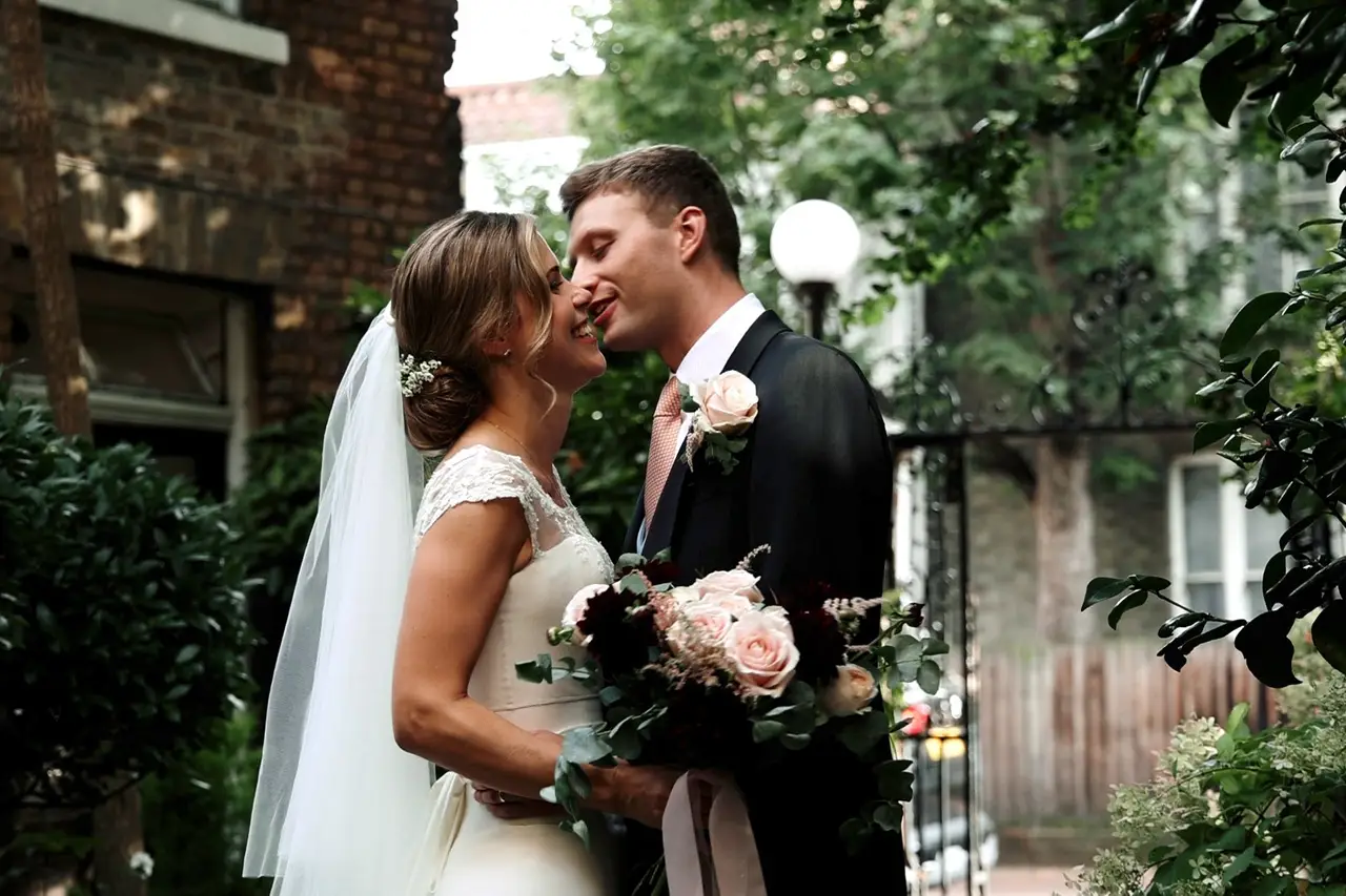 Bride holding bouquet kissing groom in front of lush greenery